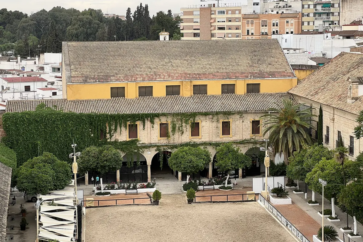 A view from above of a large stone courtyard with orange trees, an arcade, a sand arena and a yellow-walled stable range behind it, with white houses beyond