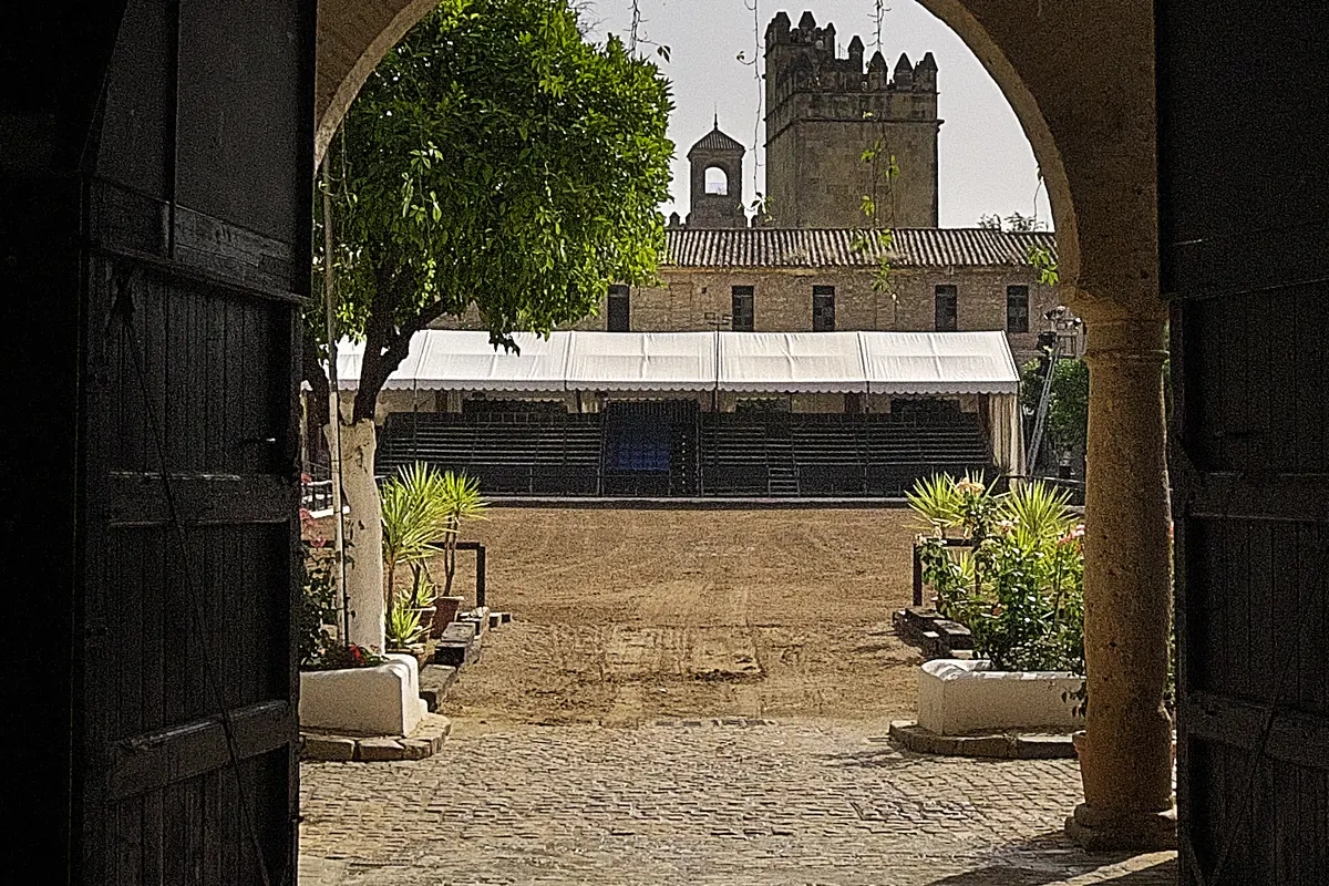 Looking through a dark wooden gate and a stone arch into a sunlit courtyard with an orange tree, potted plants, a sand arena and an old castle tower beyond