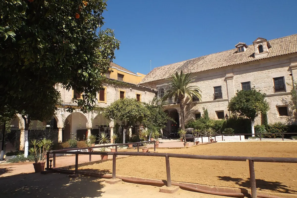 A sunlit courtyard with orange trees, a palm, a timber rail around a sand arena and a stone stable range behind it.