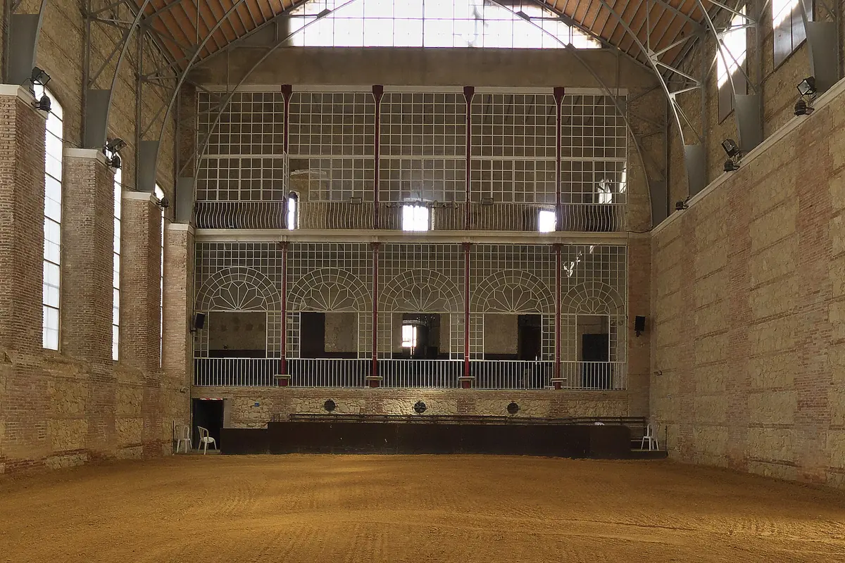 The inside of a tall covered riding hall with brick walls, high windows and a roof truss over a sand arena