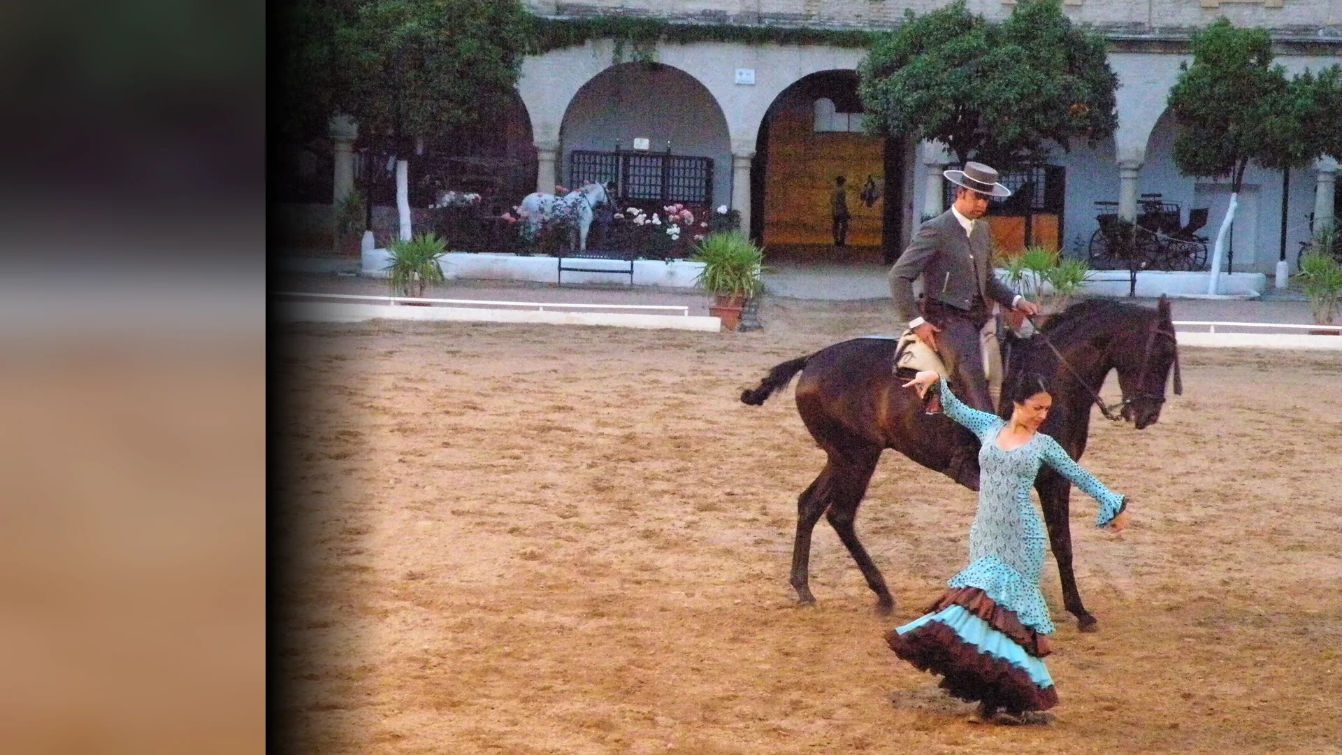 A flamenco dancer in a turquoise ruffled dress turns beside a rider on a dark bay horse in a sand arena, with white arcades and orange trees behind them.